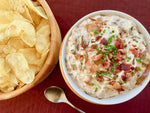 Pictured is a bowl of regular potato chips alongside a bowl of prepared bacon and onion dip garnished with crumbled fresh bacon and chopped chives. There is a silver spoon at the bottom of the photo in between the 2 bowls.